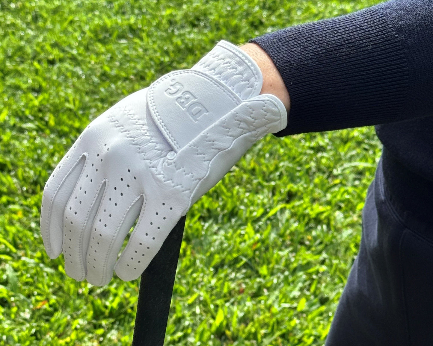 White leather golf glove on a women's hand holding a golf club with grass in the background