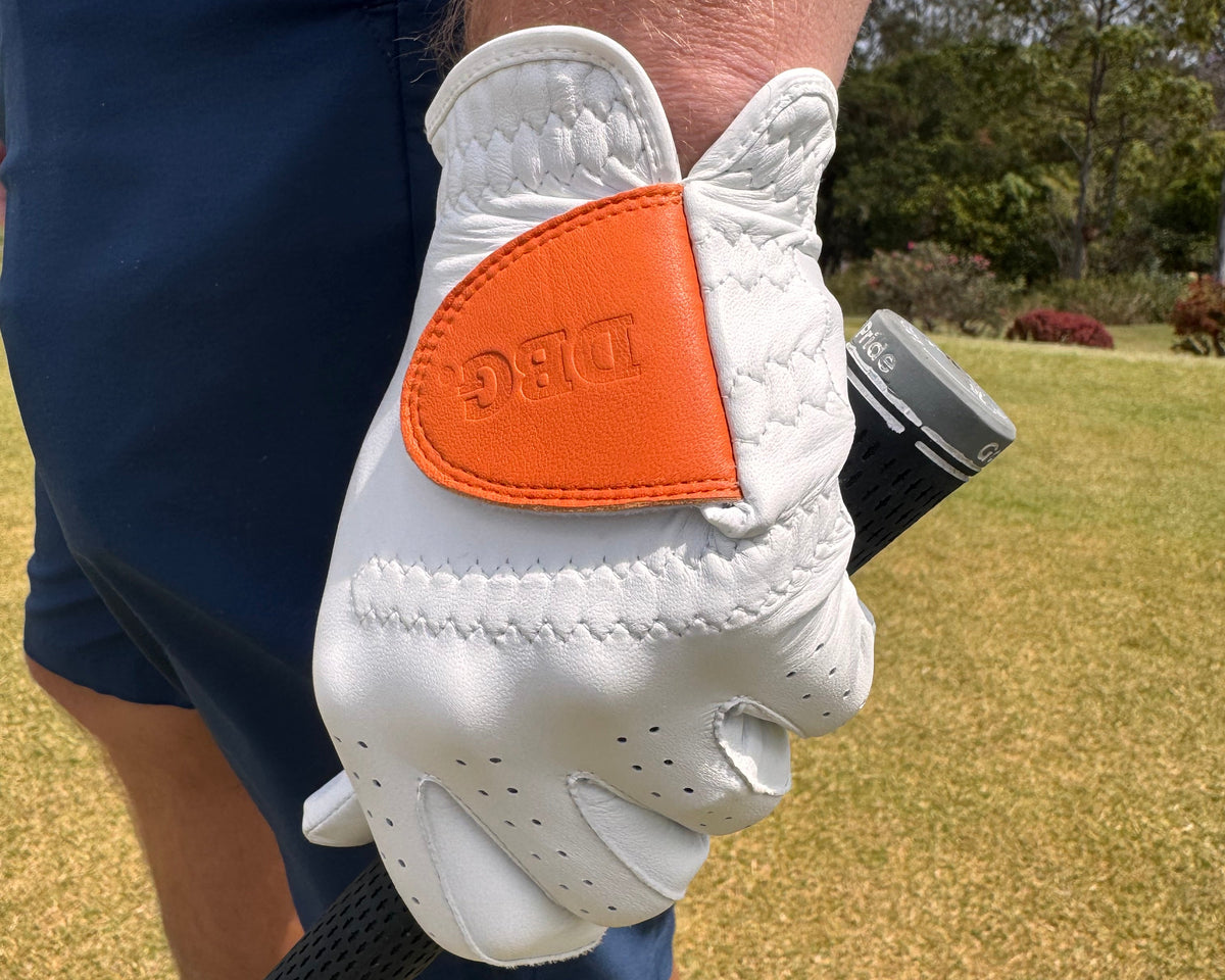 A close-up of a person's hand wearing a white golf glove with an orange accent patch and embossed 'DBG' logo, holding a golf club on a grassy golf course.