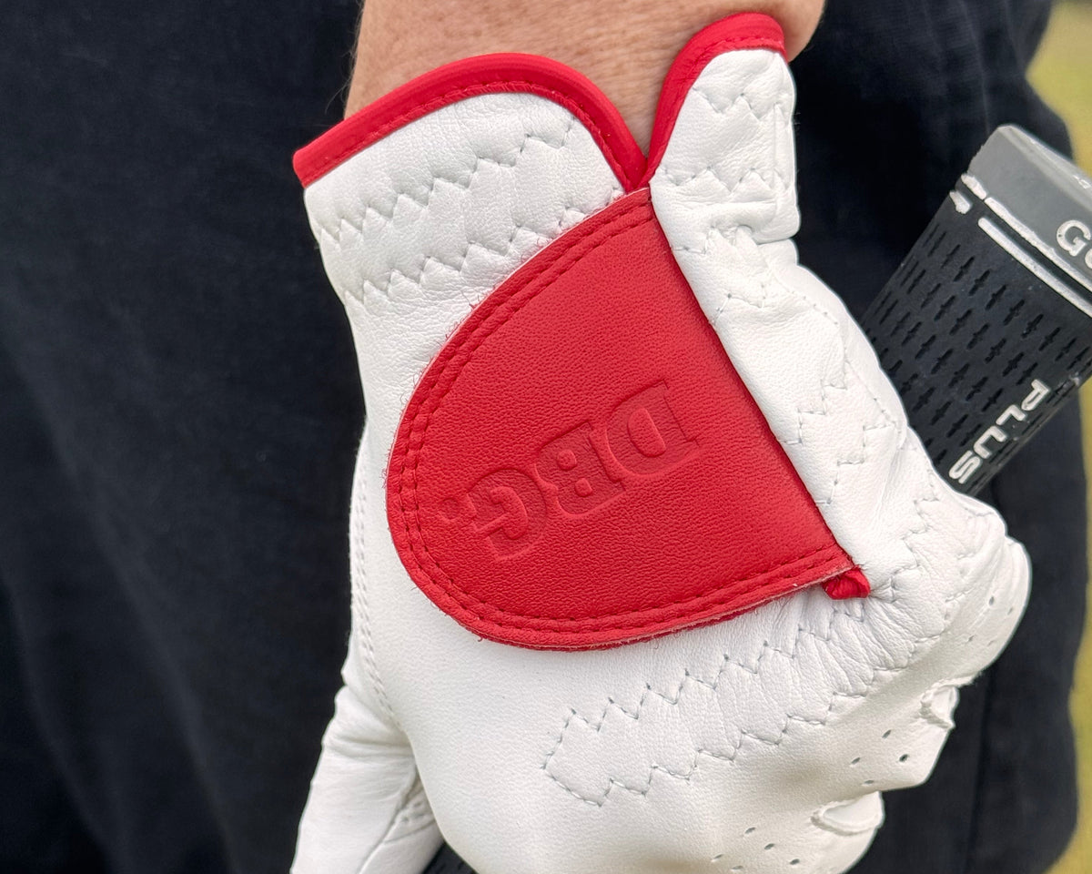A close-up of a person's hand wearing a white golf glove with a red accent panel and the debossed brand name 'DBG' on the back, holding a golf club on a golf course.
