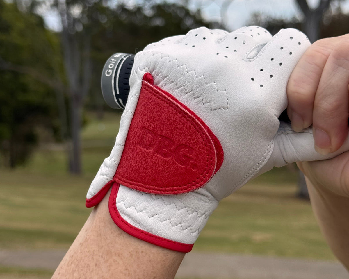A close-up of a person's hand wearing a white DBG golf glove with a red back panel, holding a golf club on an outdoor golf course.