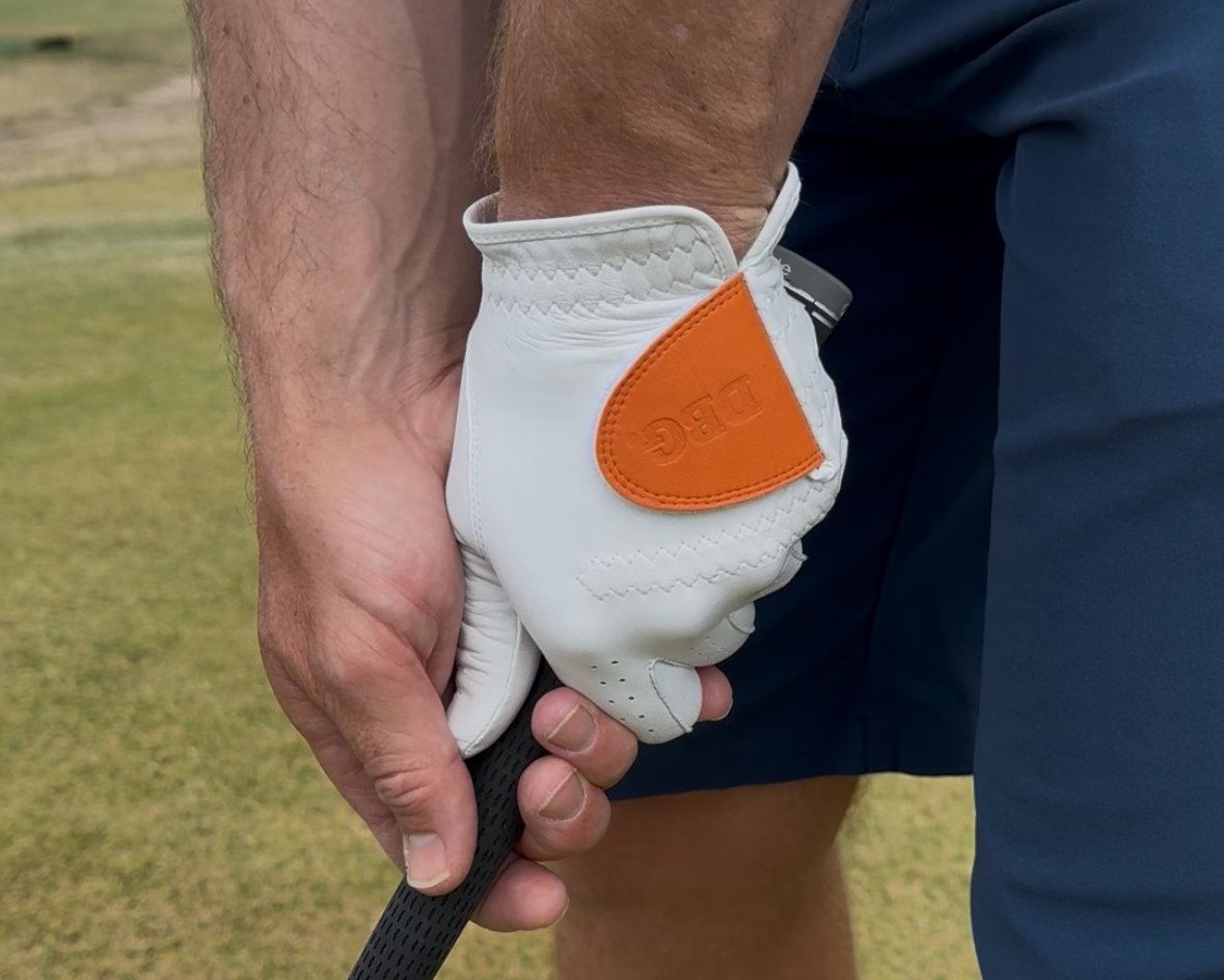 A close-up of a person's hands and legs on a golf course, wearing a white golf glove with a bright orange accent patch and holding a golf club.